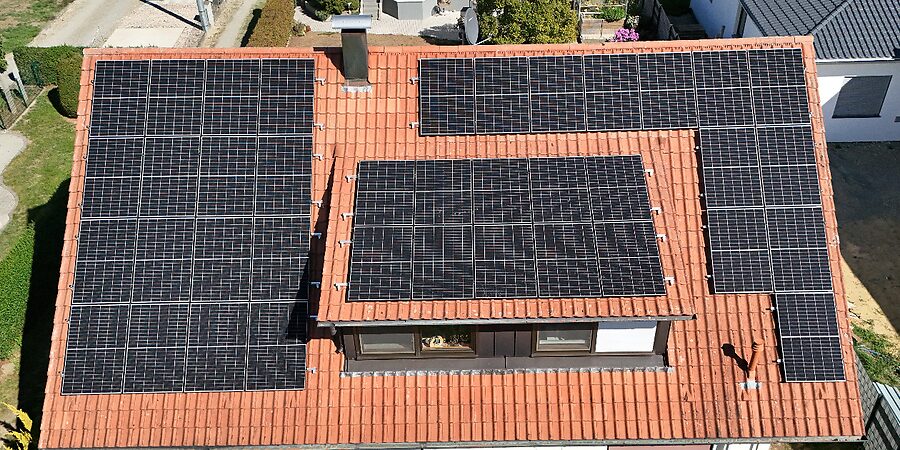 Luftaufnahme einer Photovoltaikanlage auf dem Ziegeldach eines Wohnhauses, die Module folgen dem Dachverlauf um Gaube und Kamin.